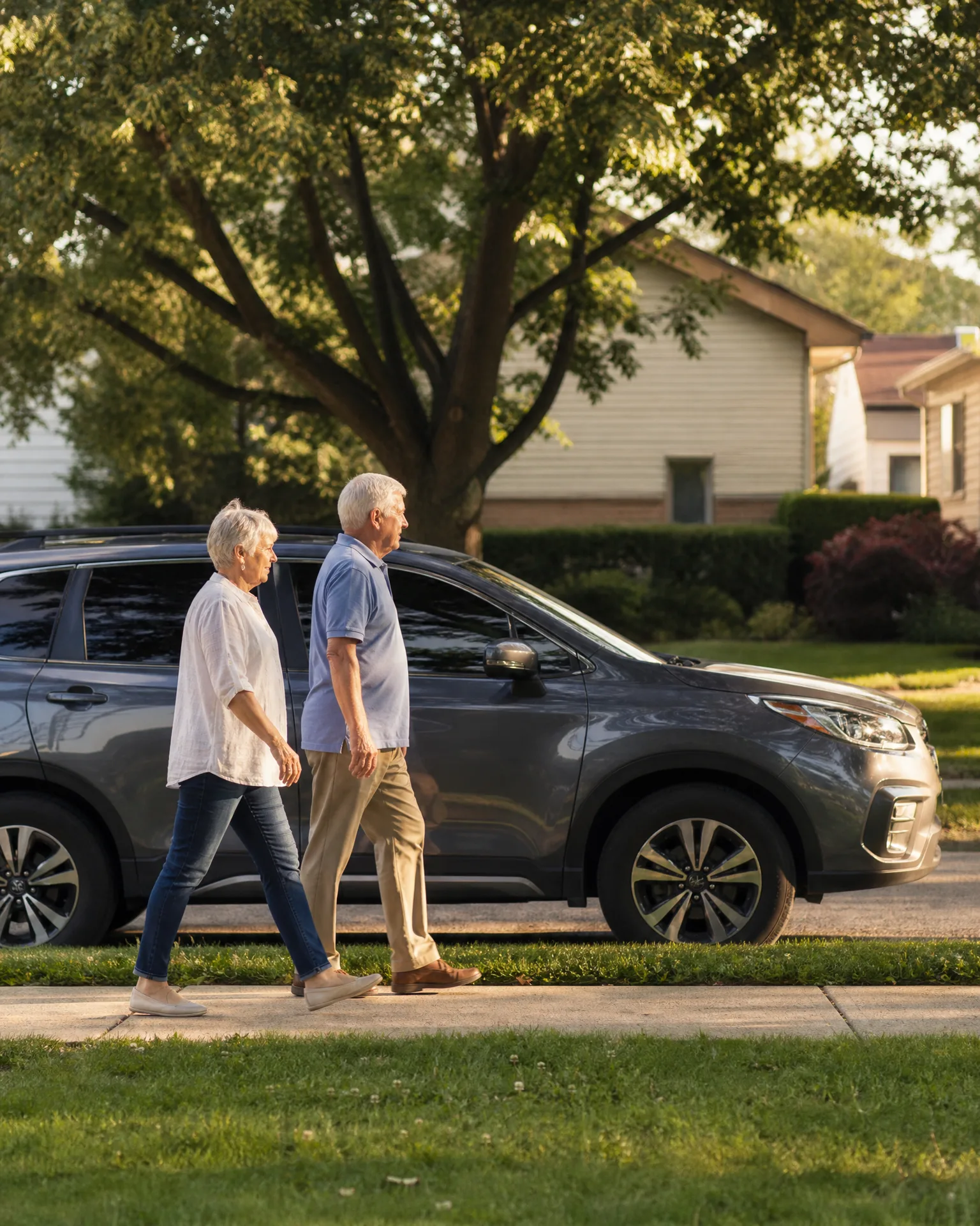 An older man and woman walk along a sidewalk past a gray SUV parked in a driveway, with a large tree and houses in the background.