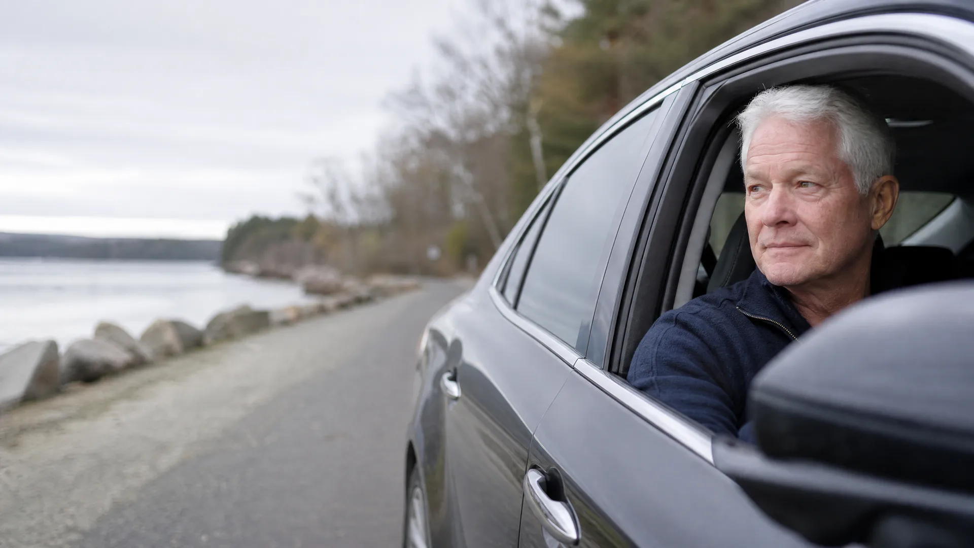 An older man with white hair leans his arm out the open driver's window of a dark car parked on a paved road beside a rocky shoreline under an overcast sky.