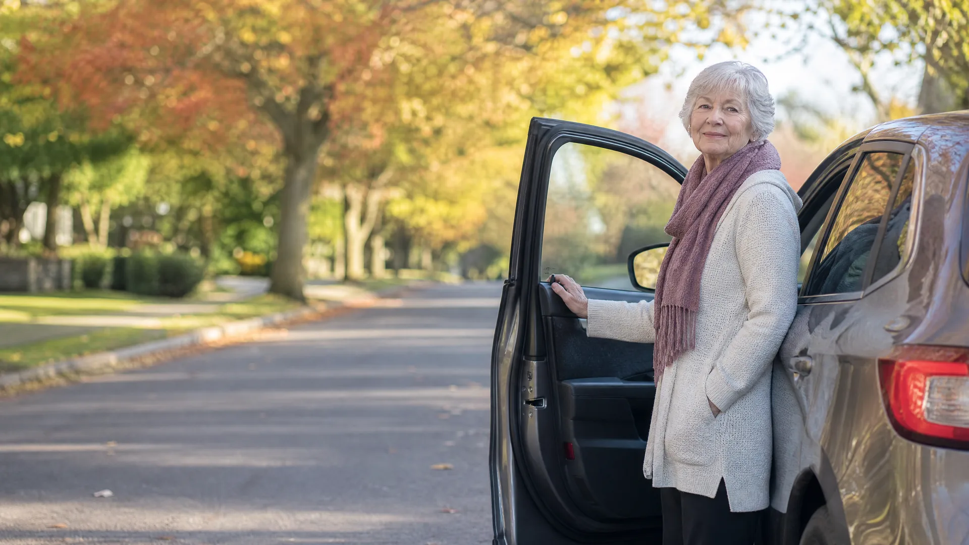 An older woman with white hair, wearing a cream cardigan and pink scarf, stands beside an open car door on a residential street lined with autumn-colored trees.