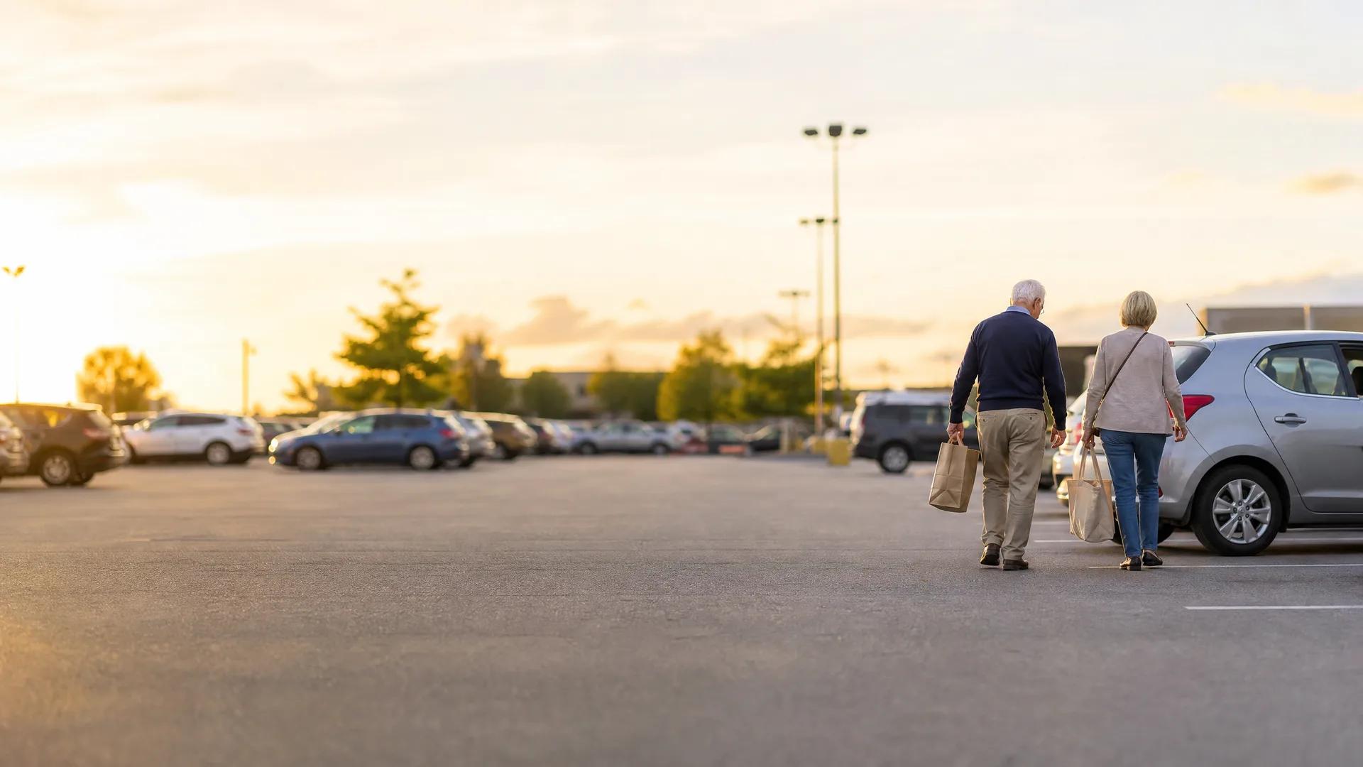 An older man and woman carrying paper shopping bags walk across a large parking lot toward a silver car at sunset, with rows of parked vehicles and a light pole in the background.