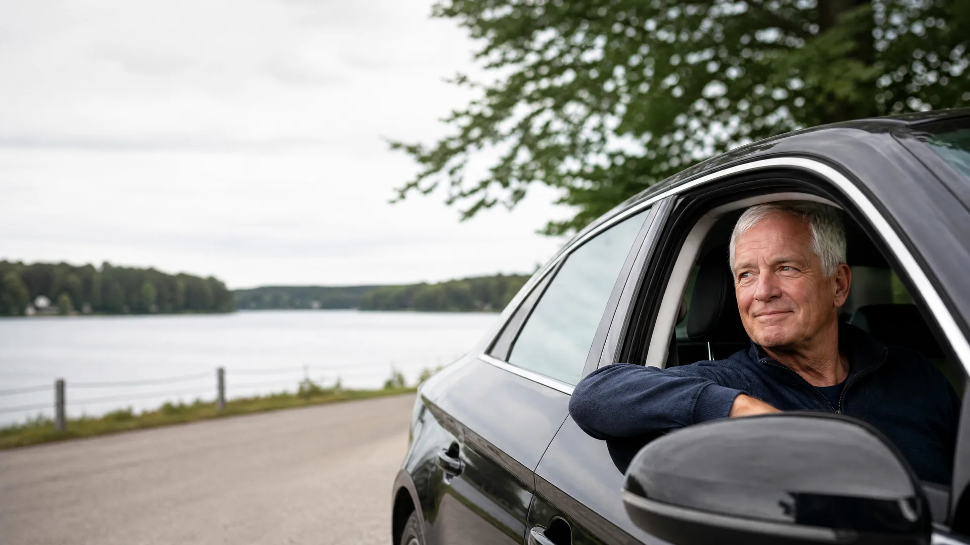 An older man with gray hair in a dark sweater leans his arm out the open driver's window of a dark car parked beside a road overlooking a lake with a tree-lined far shore under an overcast sky.