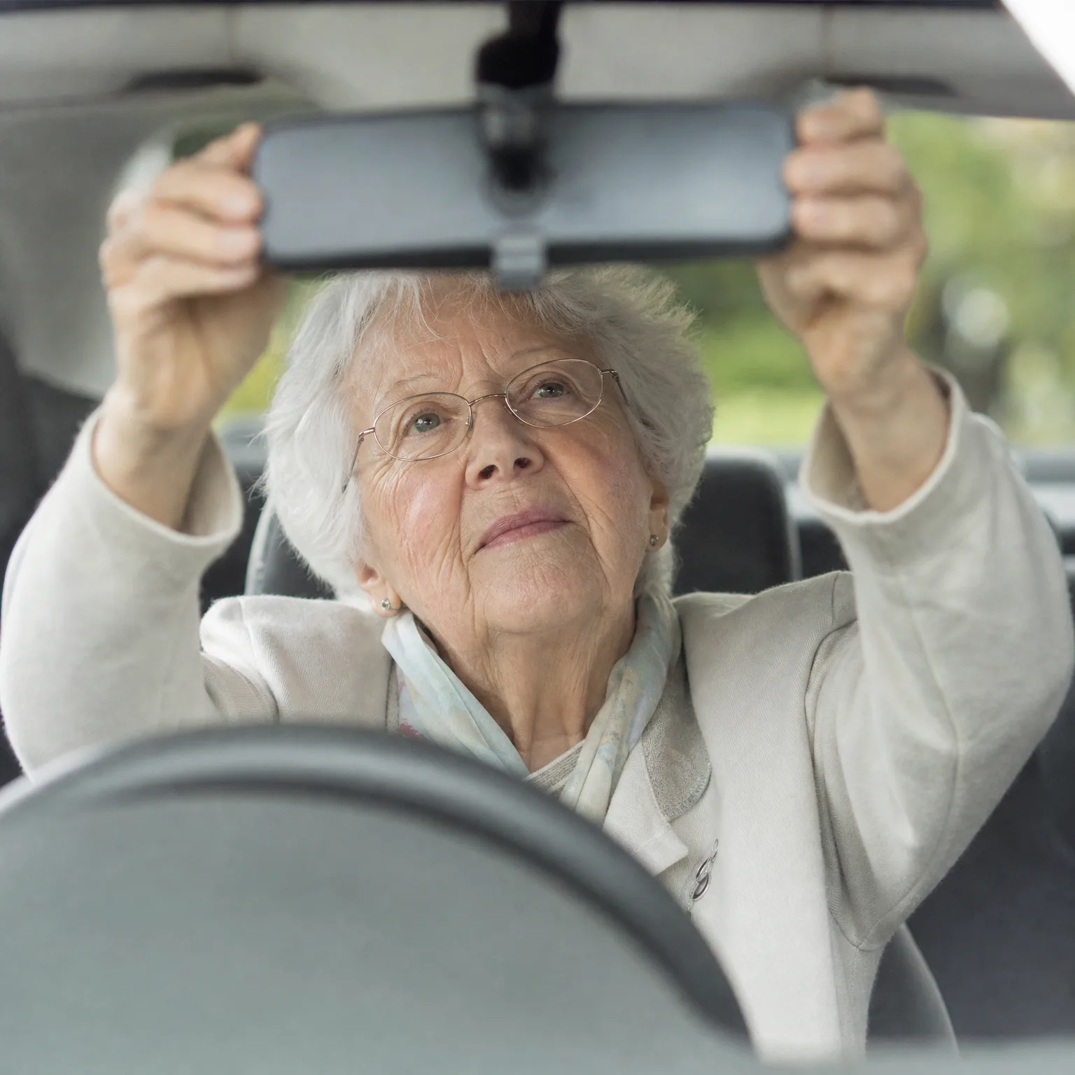 An older woman with white hair and glasses, wearing a cream jacket, reaches up with both hands to adjust a car's rearview mirror, seen from the front seat with green foliage blurred outside the windows.