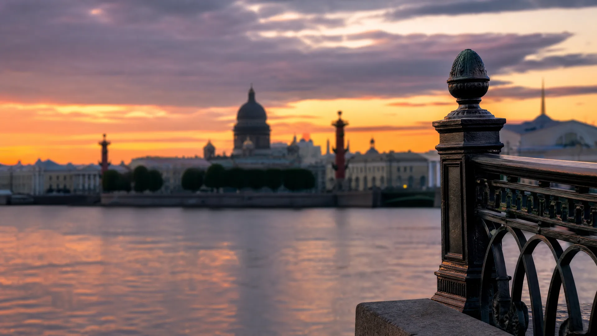 A wide river at sunset with an ornate dark metal railing and stone post in sharp focus at right, and a blurred skyline with a large domed building and columns on the far bank under orange and purple clouds.